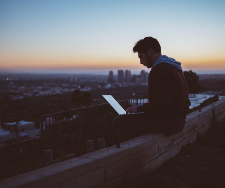 “A young man working on a laptop while seated on a ledge in a city during sunset” by Avi Richards on Unsplash.