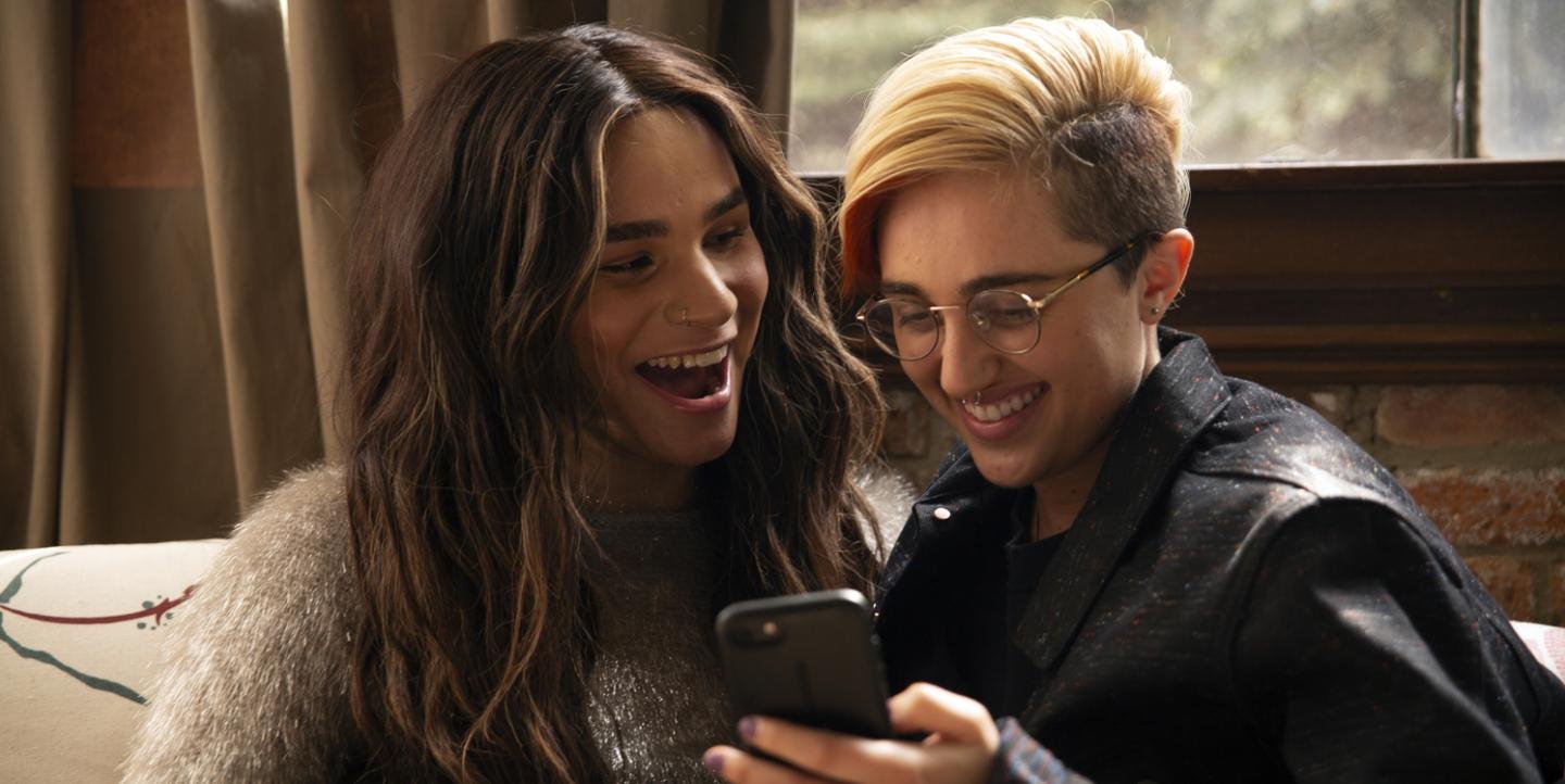 Decoration only: A photo of two non-binary or gender-neutral people, one with long brown hair and the other with shorter blonde hair, smiling as they look at someone on one of their phones.
