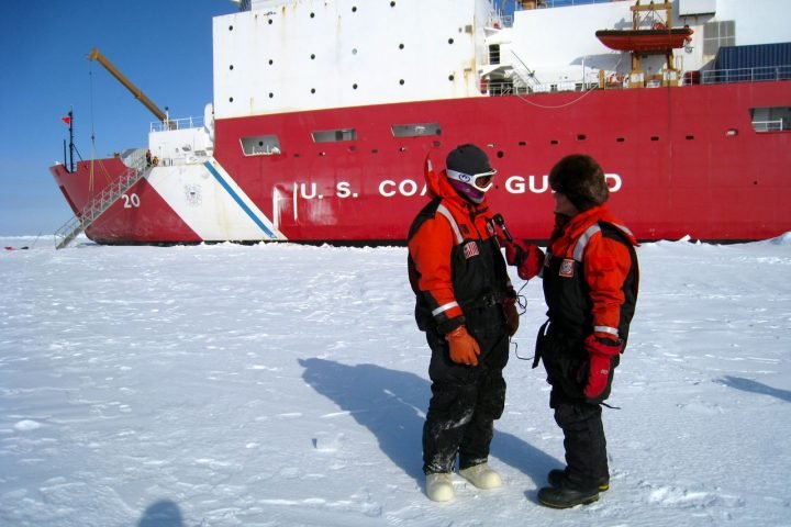 Decoration only: A photo of two U.S. Coast Guard servicemembers standing in the snow in front of a huge USCG ship breaking through the ice in what appears to be Alaska or Antartica.