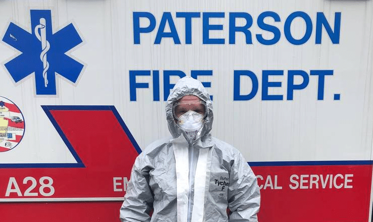A member of the Paterson Fire Department wearing a hazmat suit stands ready in front of an emergency services vehicle.