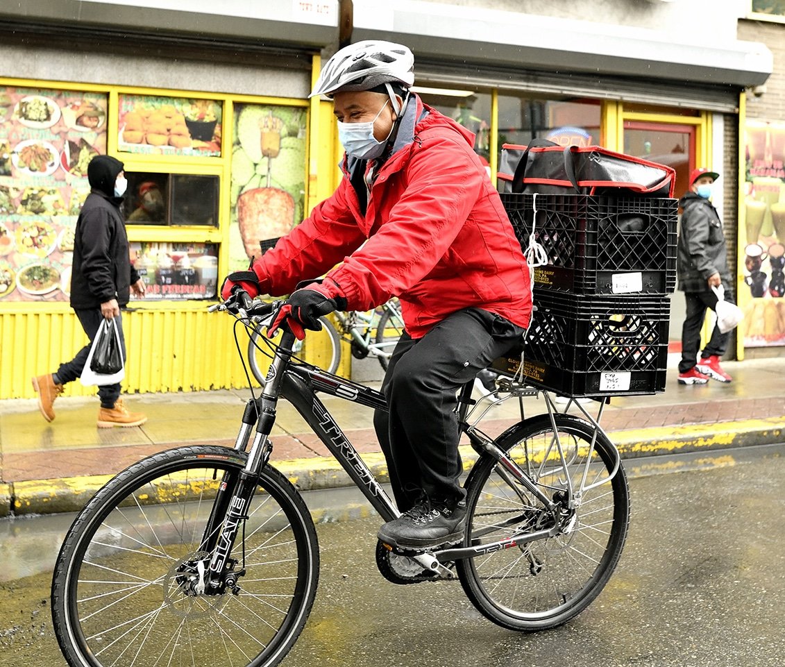 A man in a red coat and facemask delivering food via bicycle.