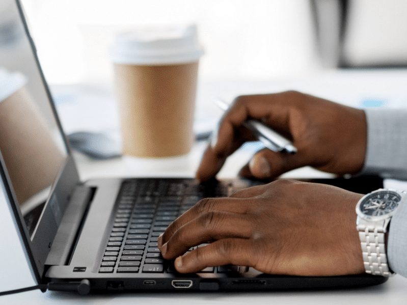 A person sits at a laptop holding a pen in hand with a coffee cup but only their hands are visible.