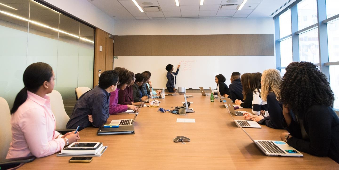 Decoration only: An instructor stands at the front of a classroom or meeting room and points at a whiteboard while other people sit at the table and listen.