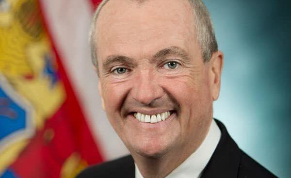 An official photo of NJ Gov. Phil Murphy standing in front of the state flag.