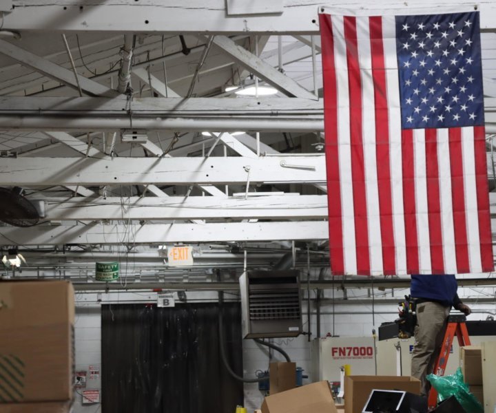 A man repairs a light on a beam behind an American flag that hangs over the factory floor. The factory is based in Paterson, New Jersey, and is manned primarily by immigrant workers.