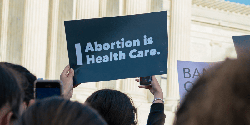Someone in a crowd of demonstrators in front of the U.S. Supreme Court holds up a blue sign with white text that reads, "Abortion is health care."